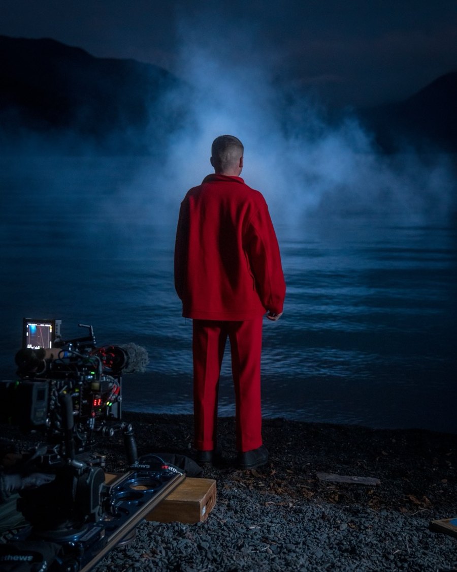 Macklemore in red standing at the edge of a misty lake at night, cinema camera rig in the foreground