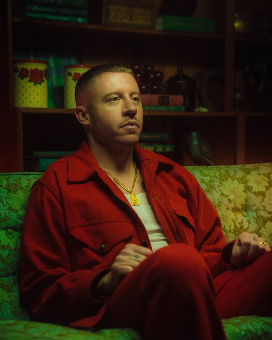 Macklemore in a red suit seated on a green floral sofa