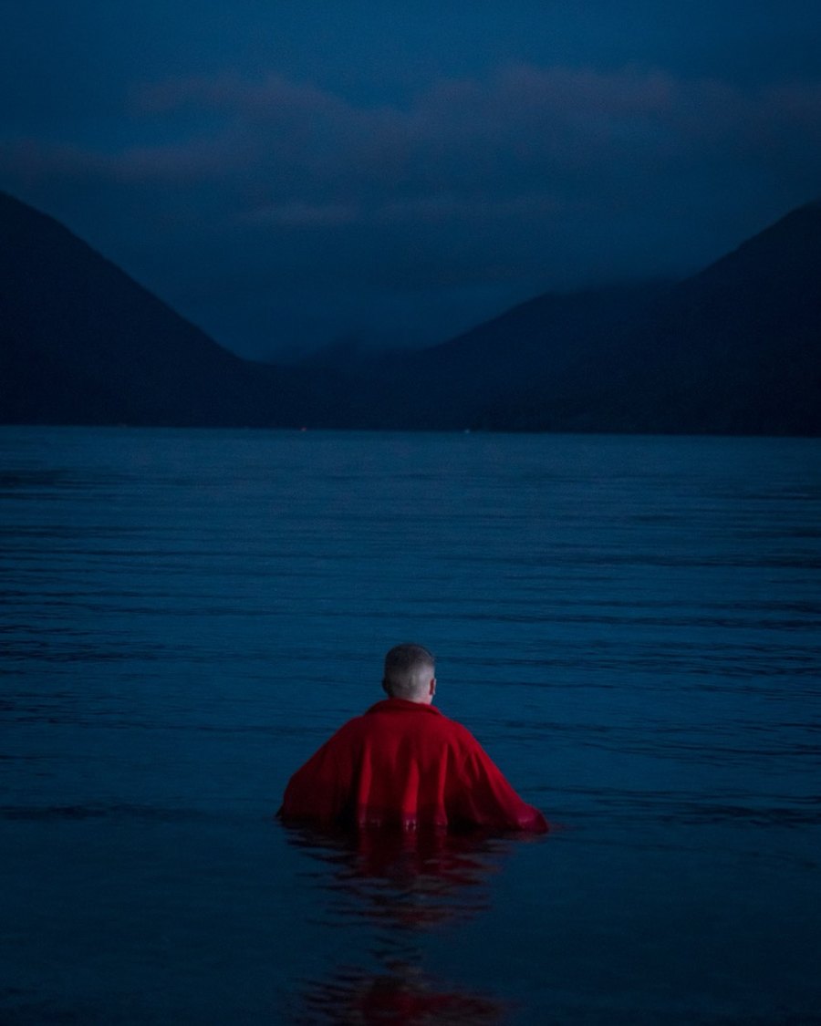 Macklemore in a red jacket wading into a dark blue lake beneath the mountains