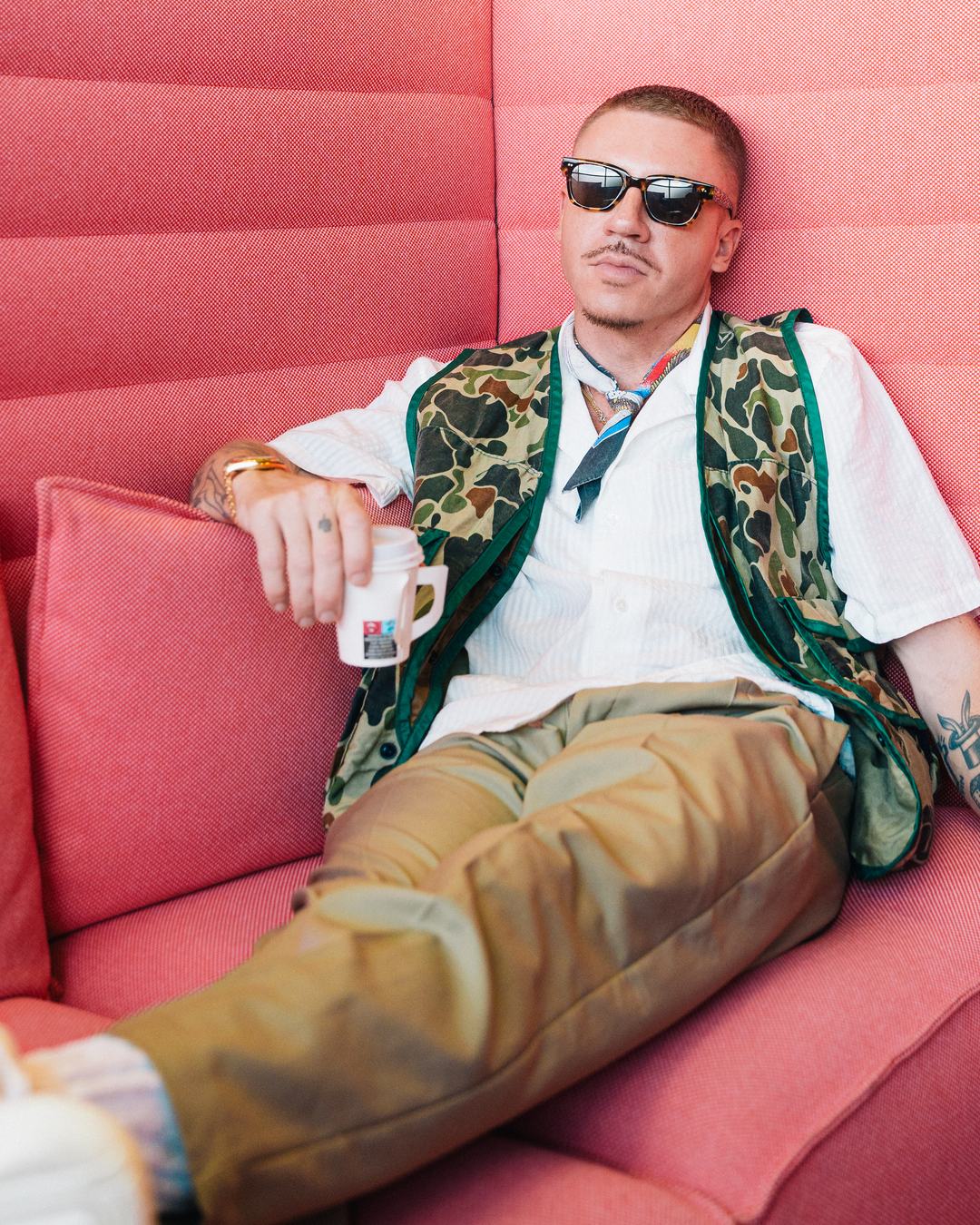 Macklemore in sunglasses and a camo vest lounging on a coral sofa with a coffee